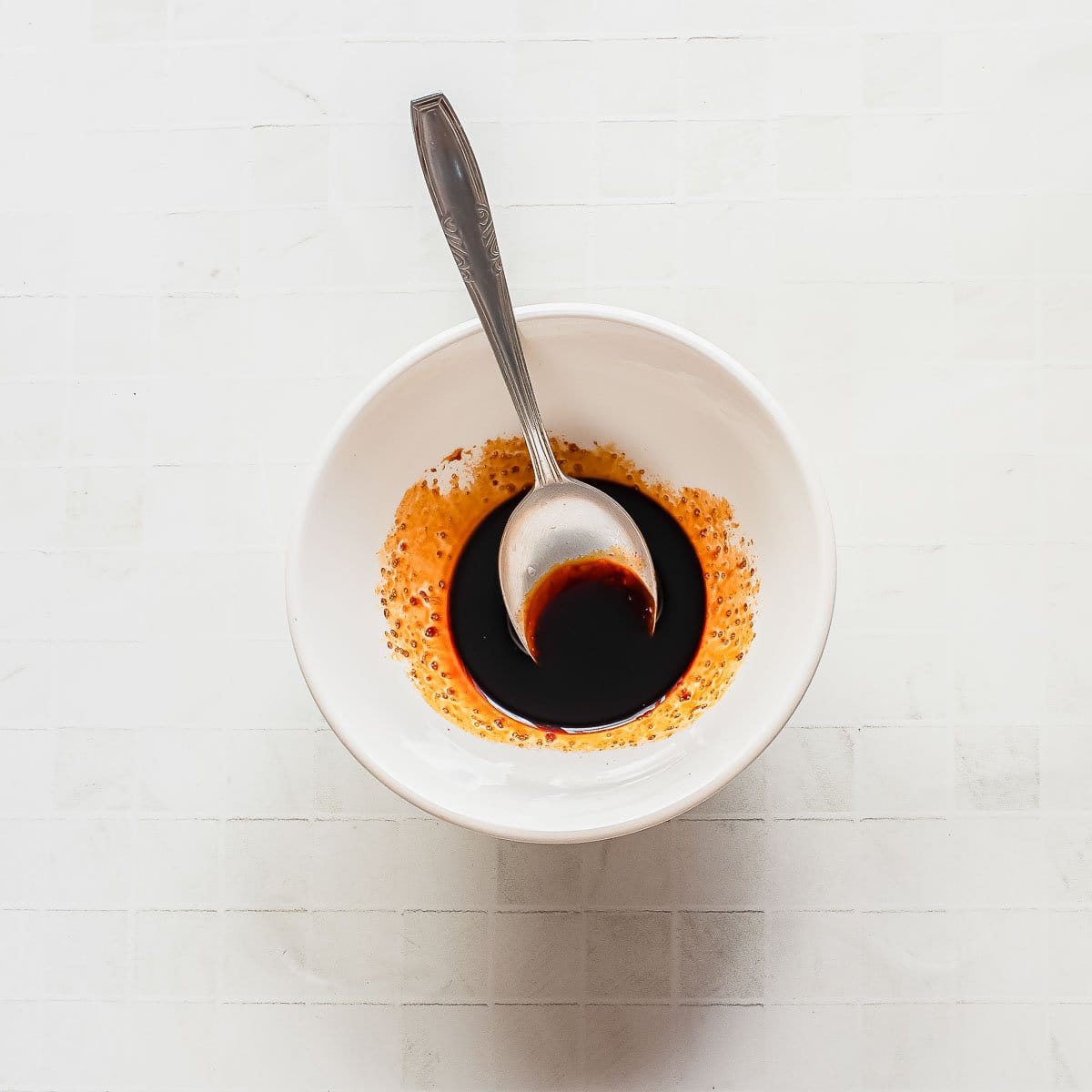 A white bowl with a metal spoon inside, containing a dark soy-based sauce with a ring of orange chili oil around the edges, sits on a light tiled surface.