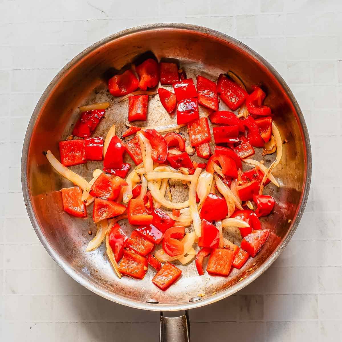 A stainless steel pan containing sautéed red bell peppers and sliced onions on a light-colored tiled surface.