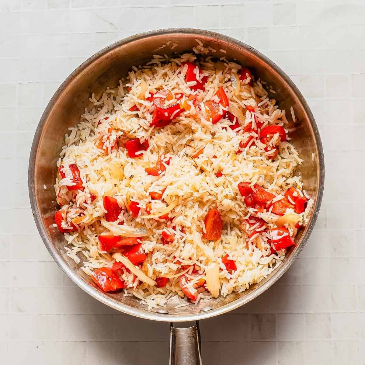 A stainless steel pan filled with cooked white rice mixed with red bell pepper pieces and sliced onions, placed on a white tiled surface.