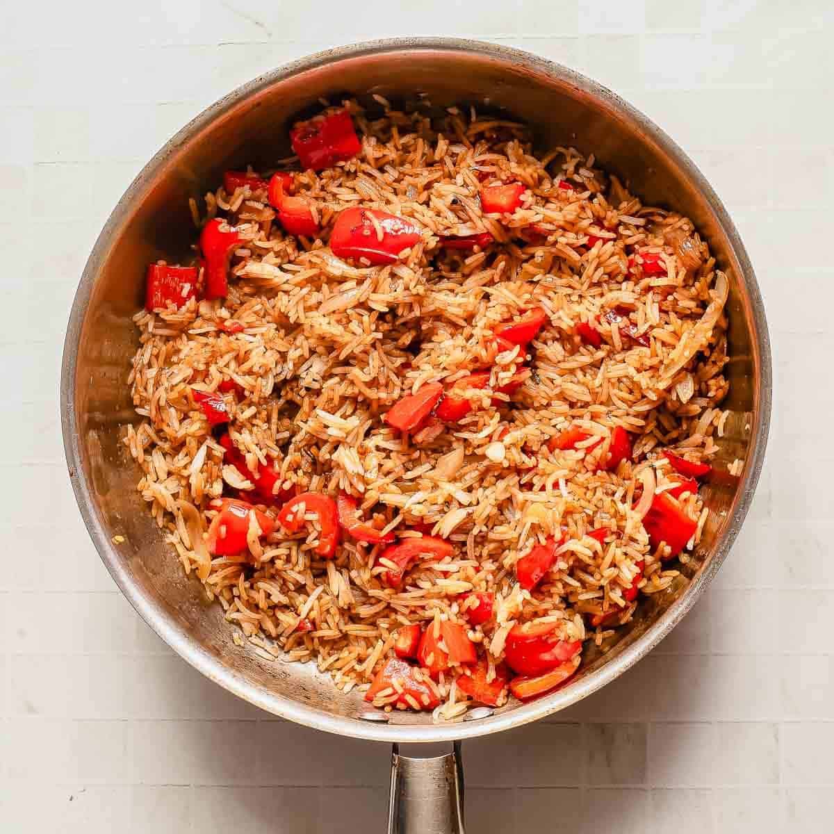 A stainless steel pan filled with cooked rice mixed with chopped red bell peppers and onions, set on a light-colored, tiled surface.