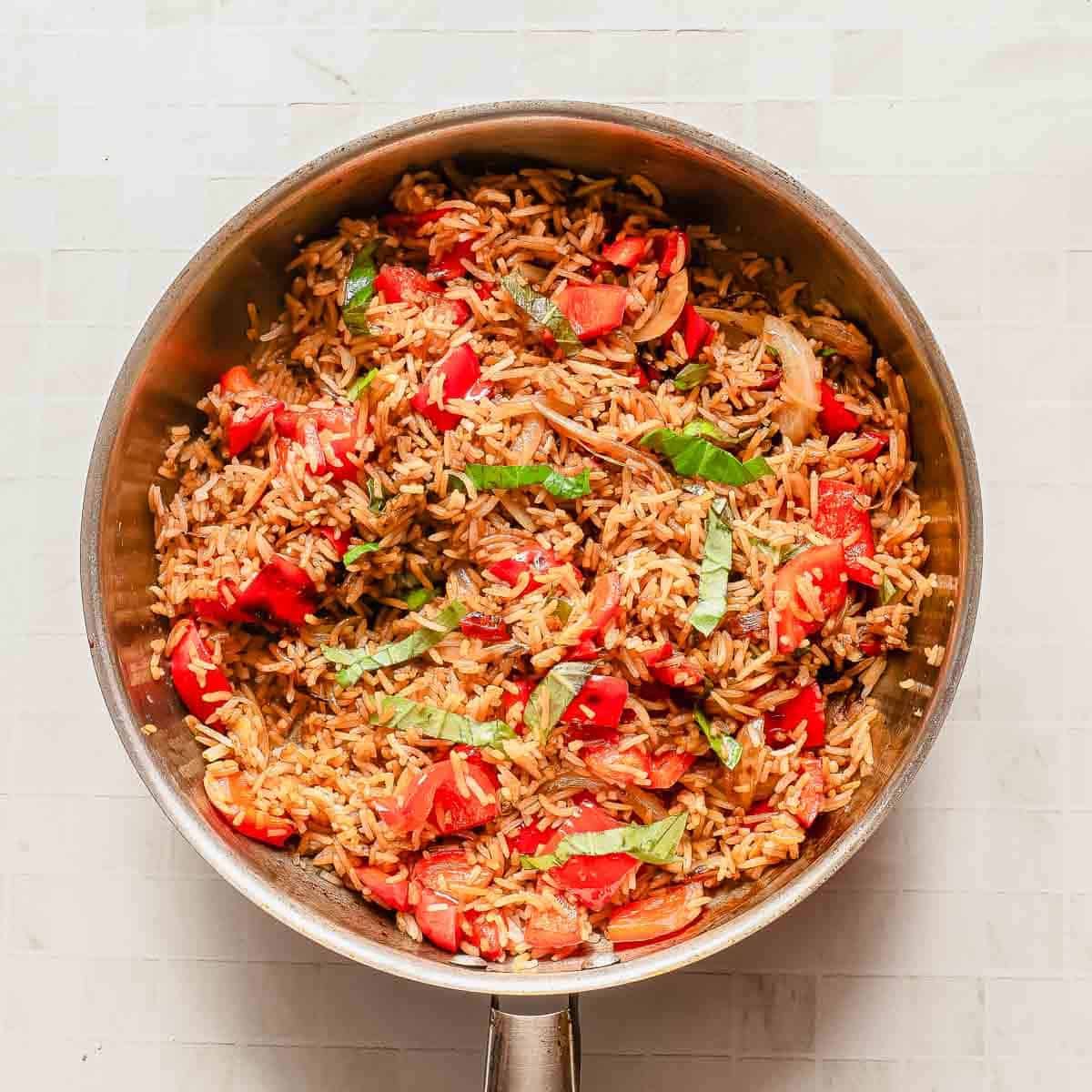 A pan filled with fried rice mixed with chopped red bell peppers, onions, and fresh basil, sitting on a light-colored tiled surface.