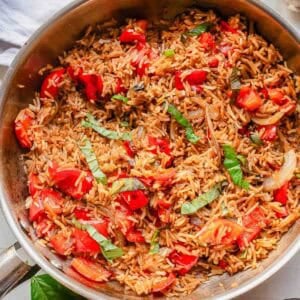 A skillet filled with basil fried rice, chopped tomatoes, sliced onions, and fresh basil strips, all mixed together and seasoned, sits on a light-colored surface with a basil leaf nearby.