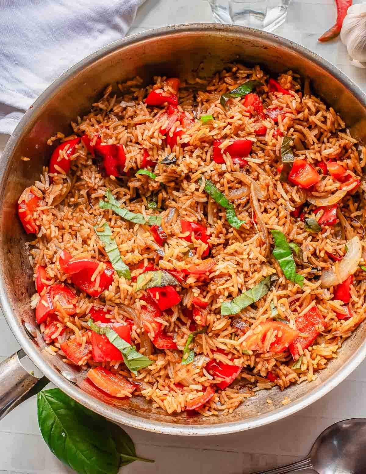 A skillet filled with basil fried rice, chopped tomatoes, sliced onions, and fresh basil strips, all mixed together and seasoned, sits on a light-colored surface with a basil leaf nearby.
