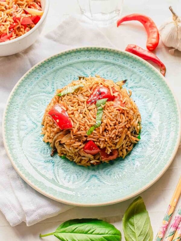 A mound of basil fried rice with red bell peppers, basil, and vegetables is served on a light blue patterned plate. Fresh basil leaves, red chili peppers, and garlic are nearby, with chopsticks and a bowl of rice in the background.