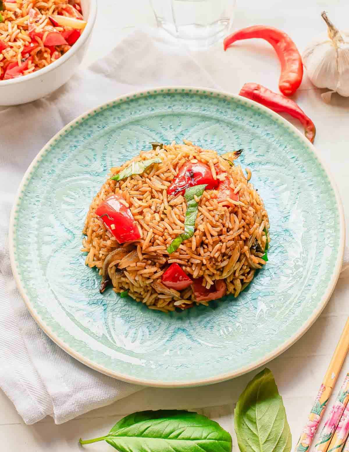 A mound of basil fried rice with red bell peppers, basil, and vegetables is served on a light blue patterned plate. Fresh basil leaves, red chili peppers, and garlic are nearby, with chopsticks and a bowl of rice in the background.