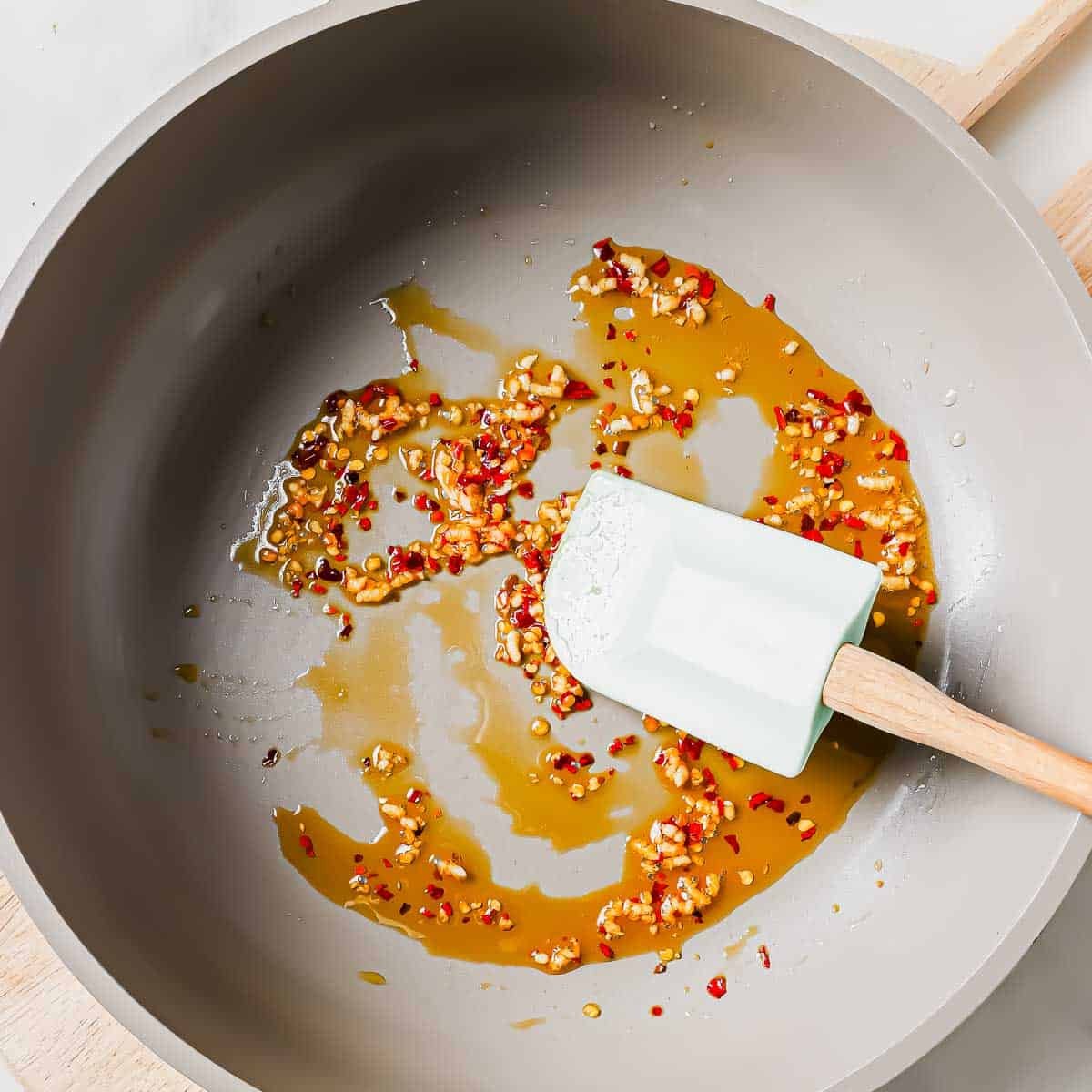 A frying pan with sautéed minced garlic and red pepper flakes in sesame oil, being stirred with a rubber spatula.