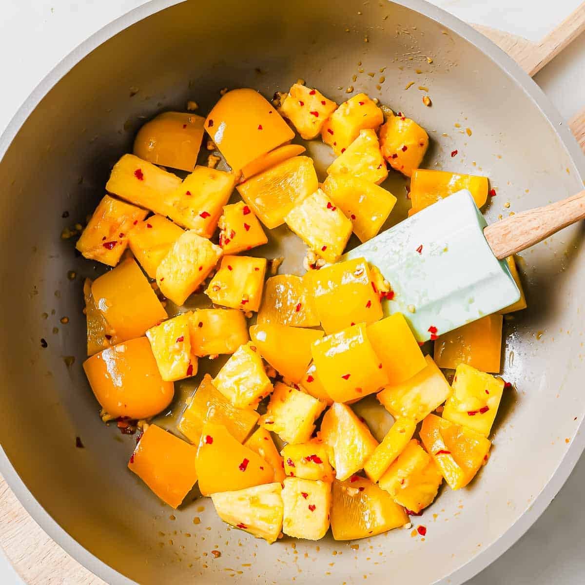 Chunks of yellow bell pepper and pineapple are being sautéed in a light-colored pan with a white spatula. The ingredients are coated in a light sauce with visible red chili flakes.
