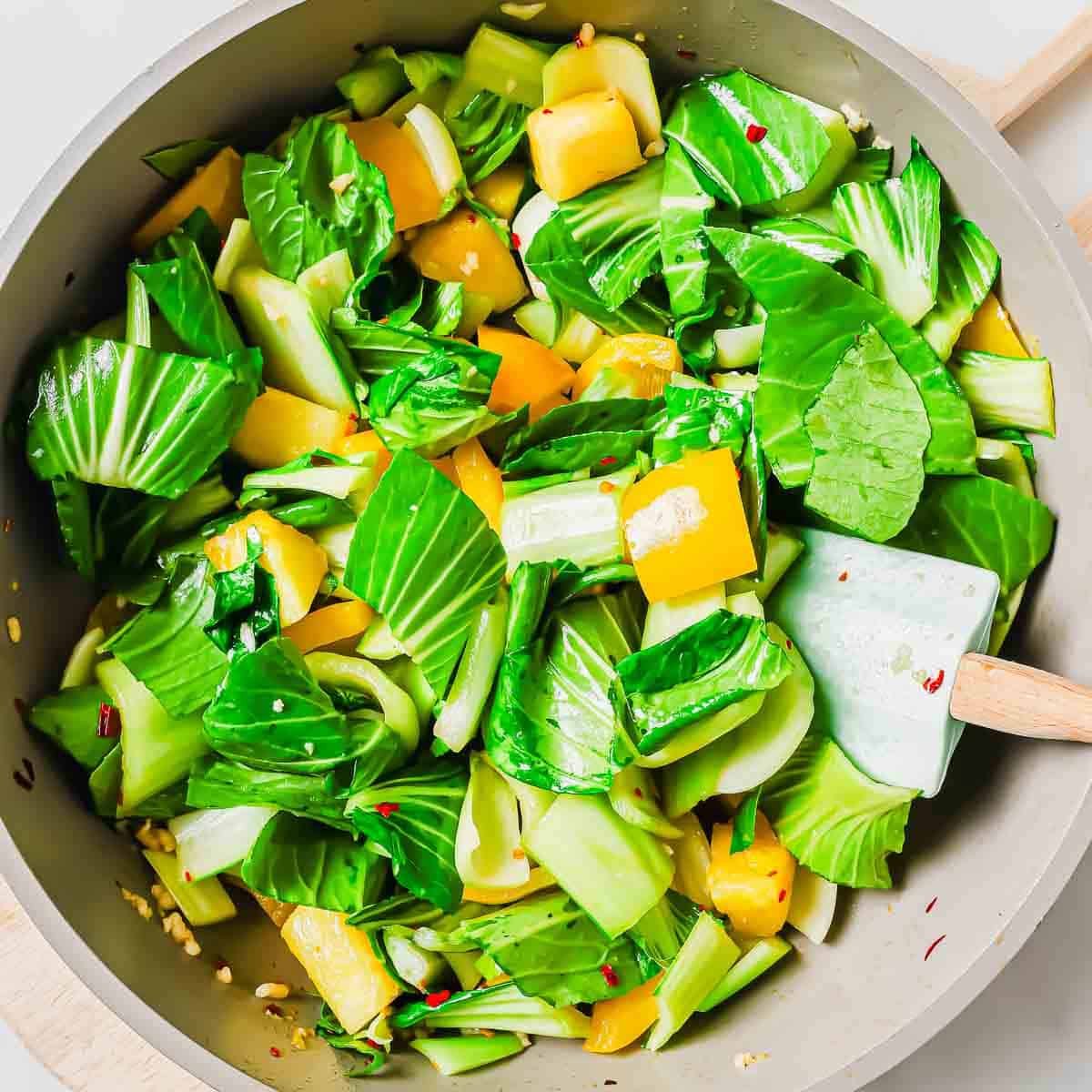 A pan filled with chopped bok choy, yellow bell pepper and pineapple being stirred with a white spatula. The vegetables are fresh, colorful, and lightly cooked.