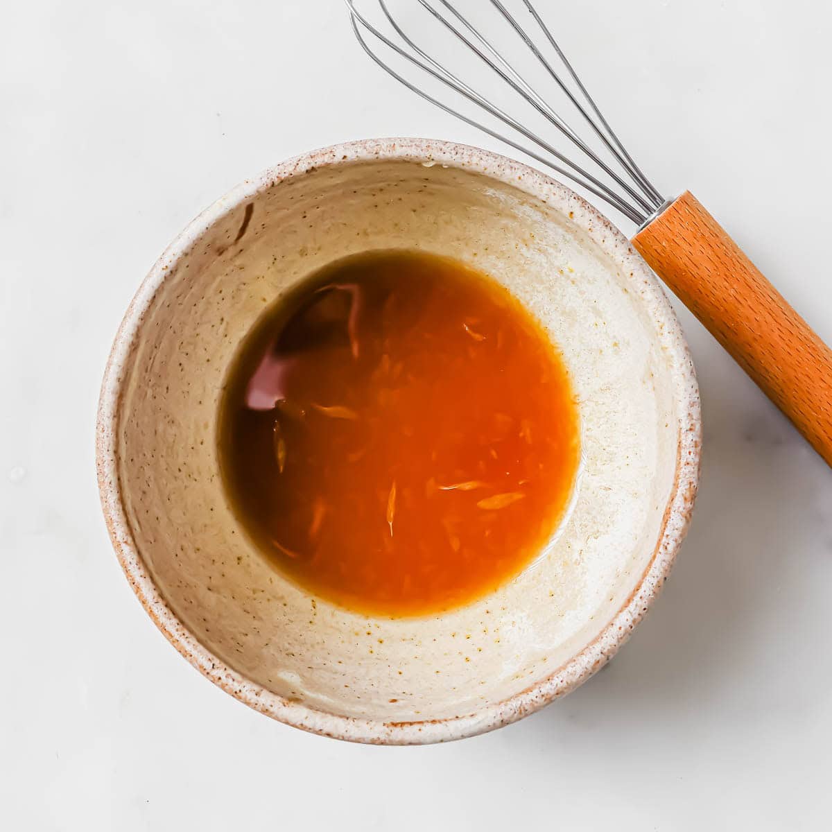 A ceramic bowl containing a soy sauce based dressing, next to a metal whisk with a wooden handle on a white surface.