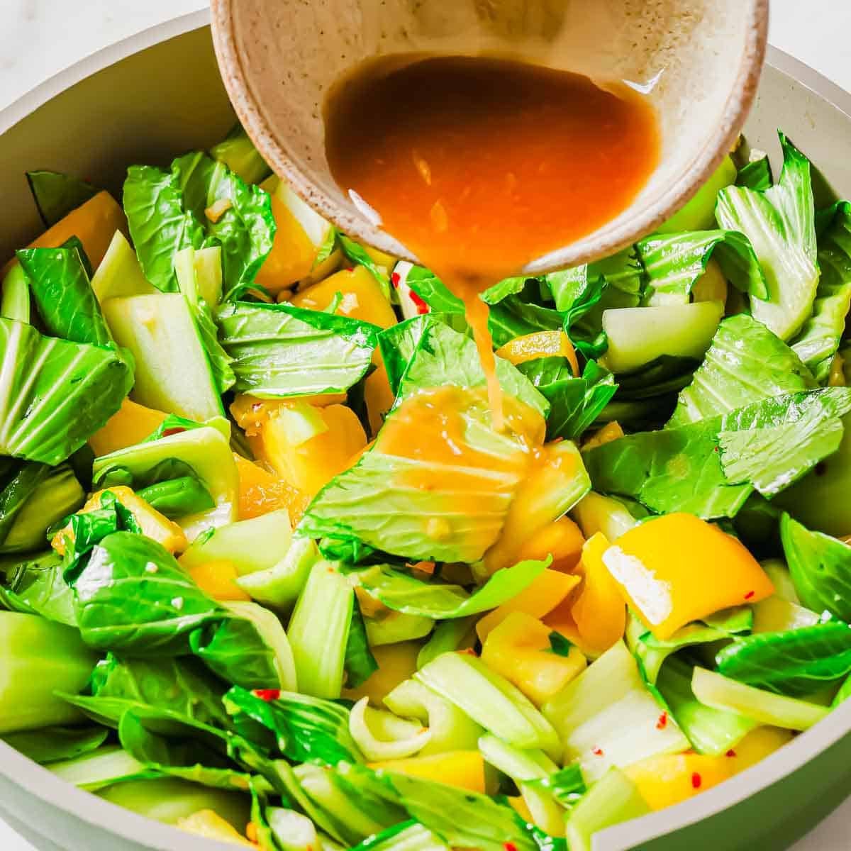 A bowl pours a brown sauce over chopped bok choy, yellow bell and pineapple peppers in a pan, preparing a colorful vegetable stir-fry.