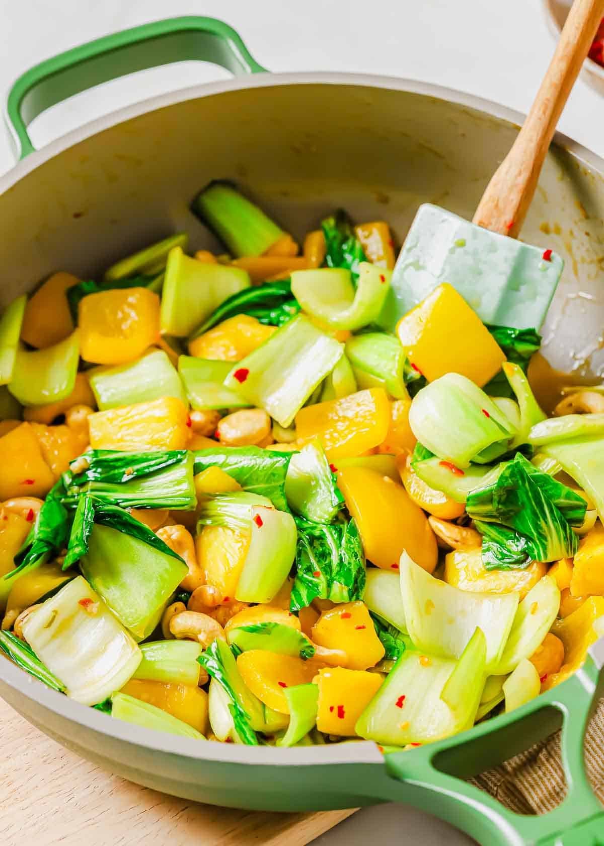 A sauté pan filled with stir-fried bok choy, yellow bell peppers, pineapple and cashews, all mixed together and being stirred with a spatula.