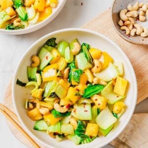A bowl of bok choy stir fry including, pineapple chunks and cashews is served on a white surface with chopsticks nearby. A small bowl of cashews and another plate of the dish are also partially visible.
