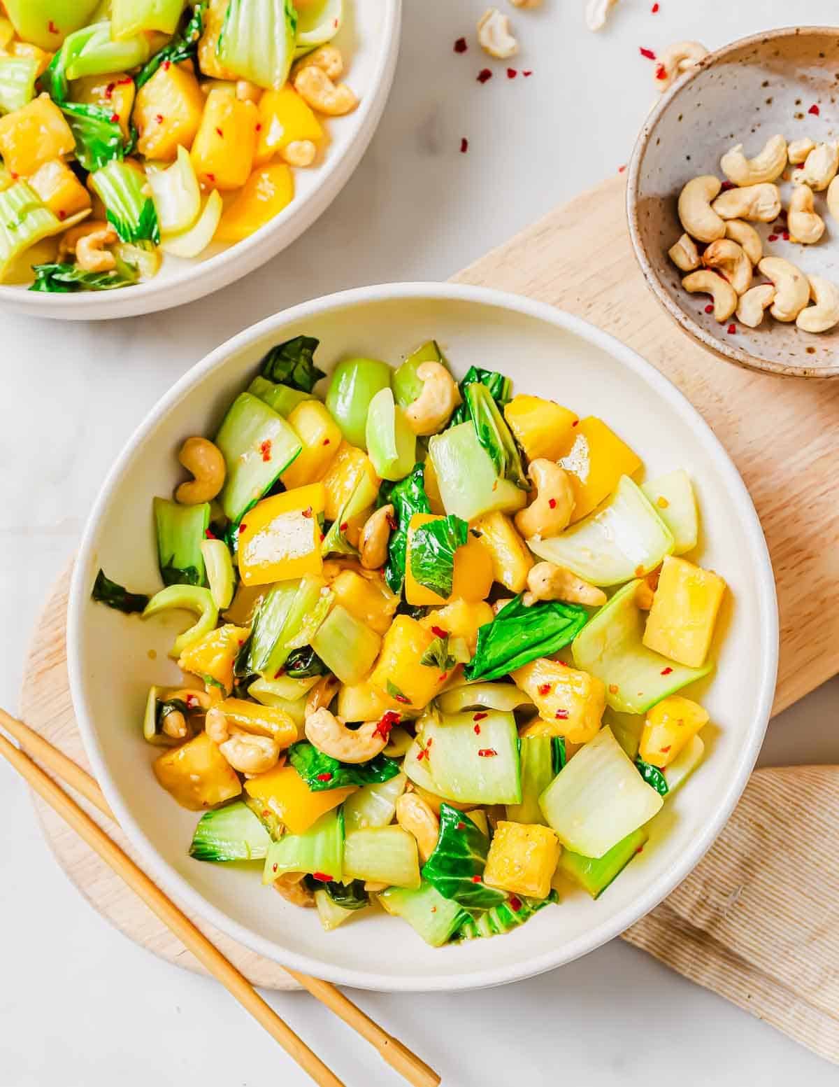 A bowl of bok choy stir fry including, pineapple chunks and cashews is served on a white surface with chopsticks nearby. A small bowl of cashews and another plate of the dish are also partially visible.