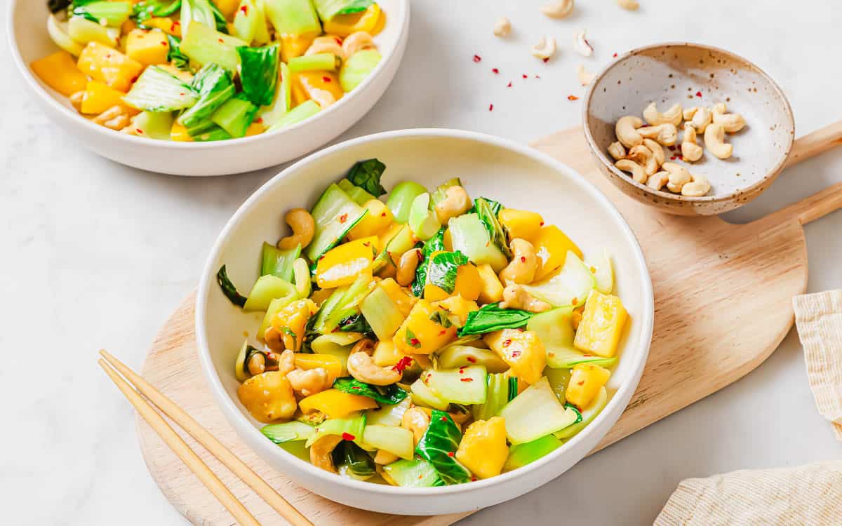 A bowl of stir-fried bok choy with pineapple, cashews, and red pepper flakes, served with chopsticks, on a wooden board. A small dish of cashews and another bowl of the dish are nearby.