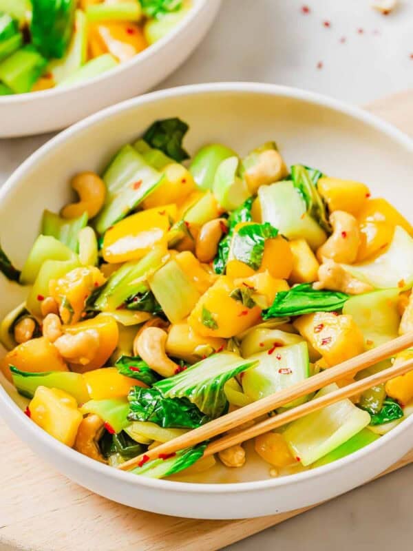 A bowl containing a bok choy stir fry with yellow bell peppers, and cashews garnished with red pepper flakes, with chopsticks resting on the side. The dish is colorful and served on a light wooden surface.