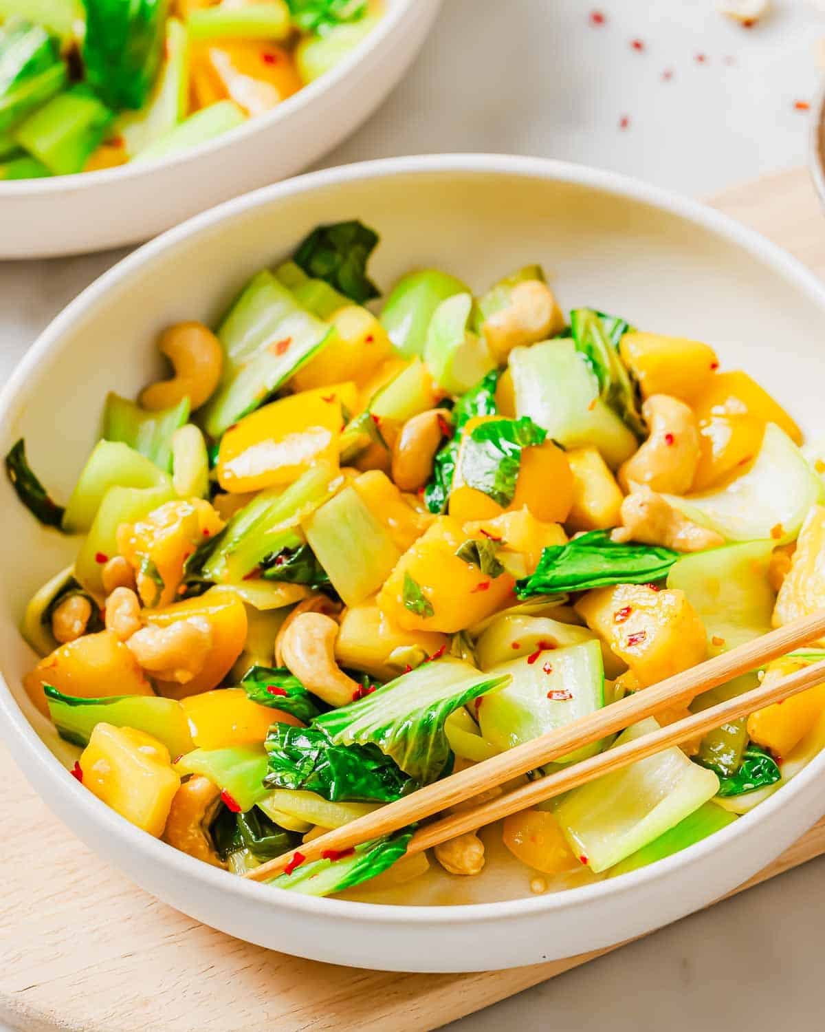A bowl containing a bok choy stir fry with yellow bell peppers, and cashews garnished with red pepper flakes, with chopsticks resting on the side. The dish is colorful and served on a light wooden surface.