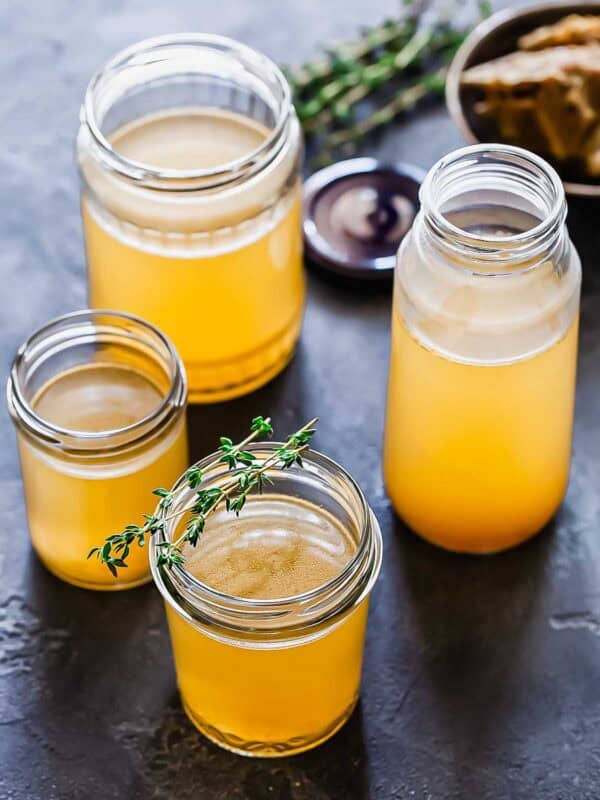 Four glass jars filled with golden bone broth are arranged on a dark surface, with one jar garnished with fresh thyme. A bowl of bones appears in the background.