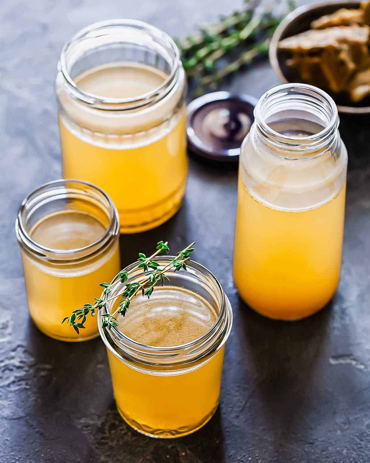 Four glass jars filled with golden bone broth are arranged on a dark surface, with one jar garnished with fresh thyme. A bowl of bones appears in the background.
