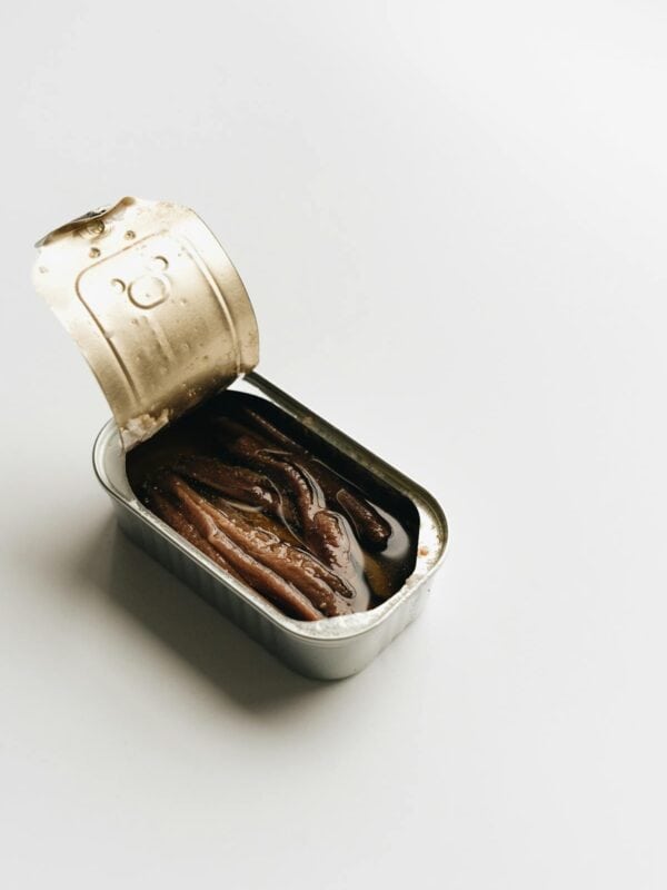 An opened tin can of anchovies in oil sits on a white surface, with the lid partially peeled back to reveal the fish inside.