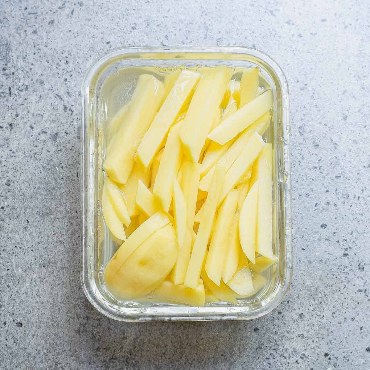 A glass container filled with raw potato slices soaking in water, placed on a gray speckled countertop.