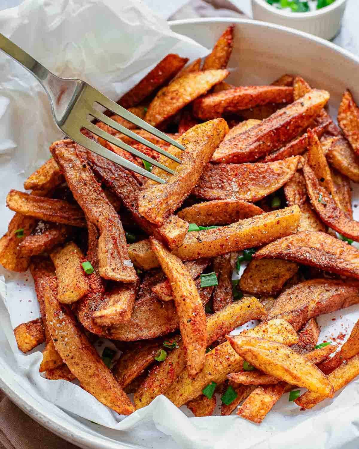 A plate of crispy, seasoned homemade masala fries garnished with chopped herbs, with a fork picking up a few pieces. The fries rest on white parchment paper inside a round dish.