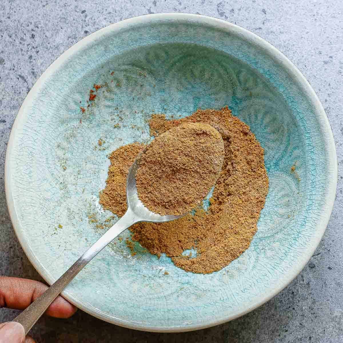 A hand holding a spoon with masala spices over a light blue bowl containing more of the same spice, on a gray countertop.