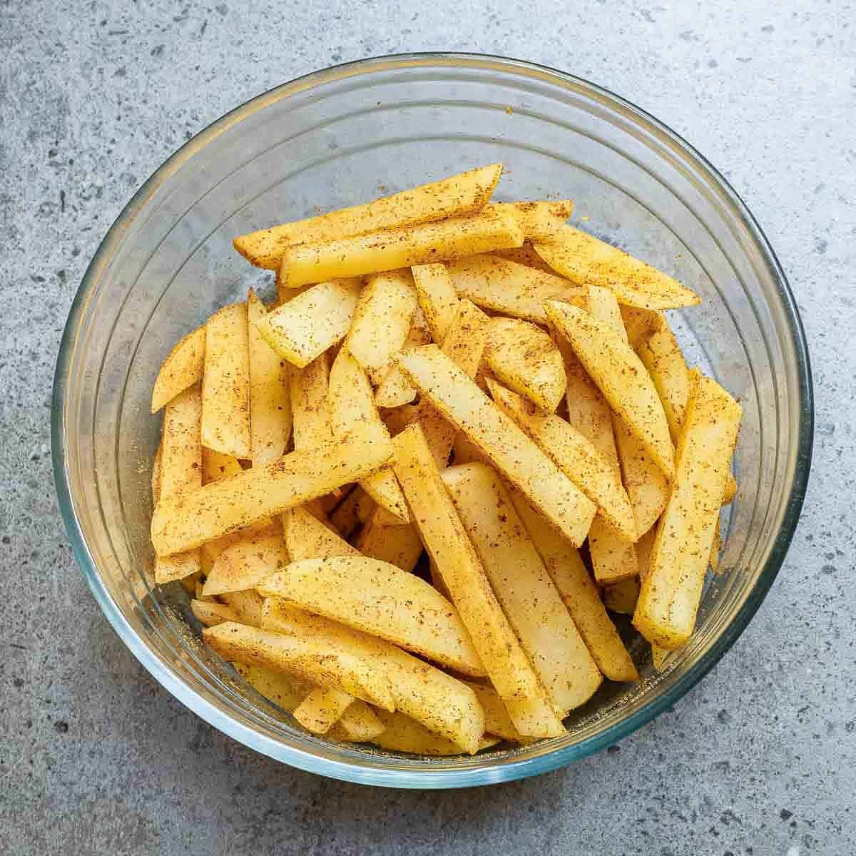 A clear glass bowl filled with uncooked potato fries coated in seasoning, placed on a gray speckled countertop.