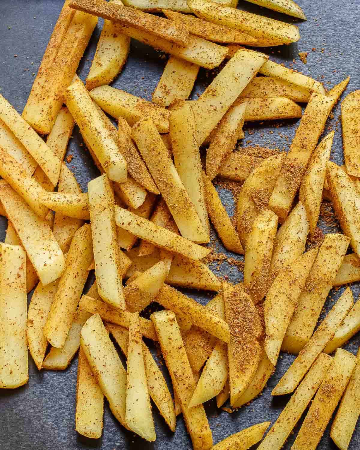 Raw potato fries sprinkled with a generous amount of masala seasoning, spread out on a dark baking tray, ready to be cooked.