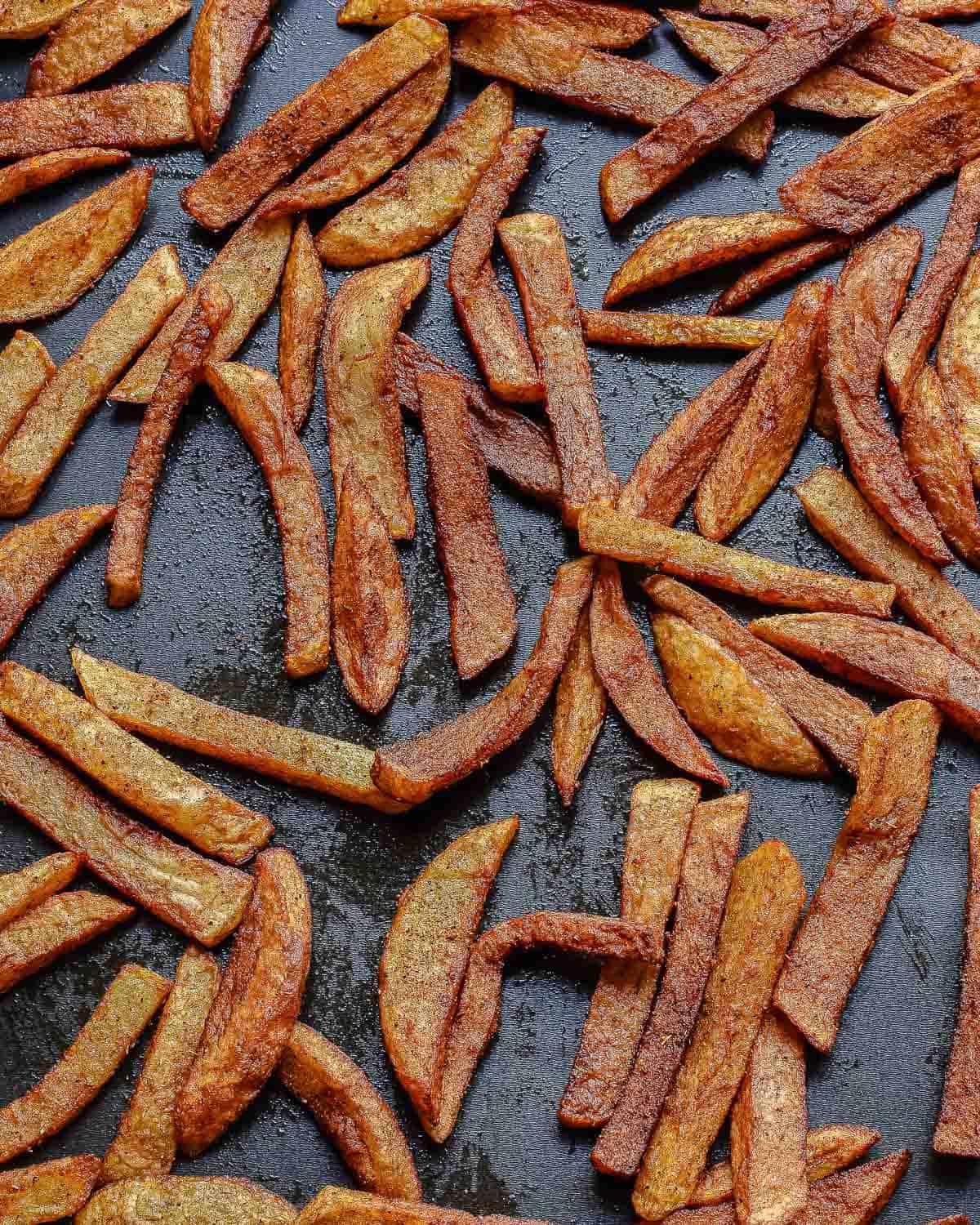 Golden-brown fries scattered on a dark, oily baking tray, showing crispy, textured edges and varying sizes, freshly baked and ready to eat.
