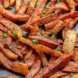 A close-up of crispy, masala spiced French fries garnished with chopped green onions, arranged on a dark, textured surface.