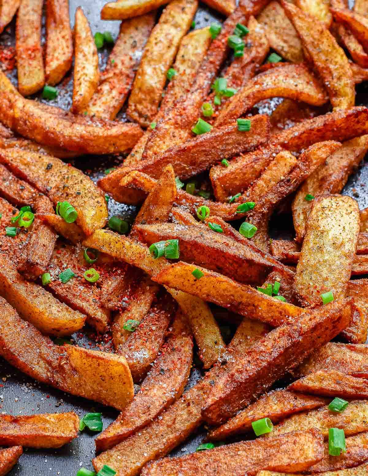 A close-up of crispy, masala spiced French fries garnished with chopped green onions, arranged on a dark, textured surface.