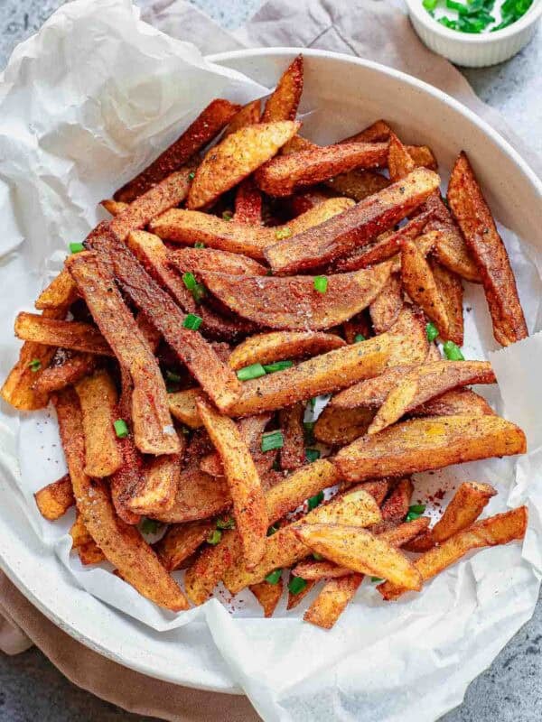 A white dish lined with parchment paper holds a pile of golden-brown, masala French fries, garnished with chopped green herbs. A small bowl with more herbs sits in the background.