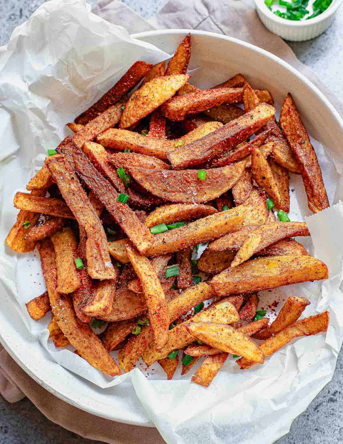 A white dish lined with parchment paper holds a pile of golden-brown, masala French fries, garnished with chopped green herbs. A small bowl with more herbs sits in the background.