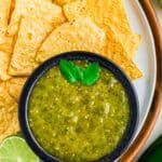 A black bowl filled with salsa verde garnished with cilantro, surrounded by yellow tortilla chips on a white plate. A lime slice and part of a jalapeño are also visible.