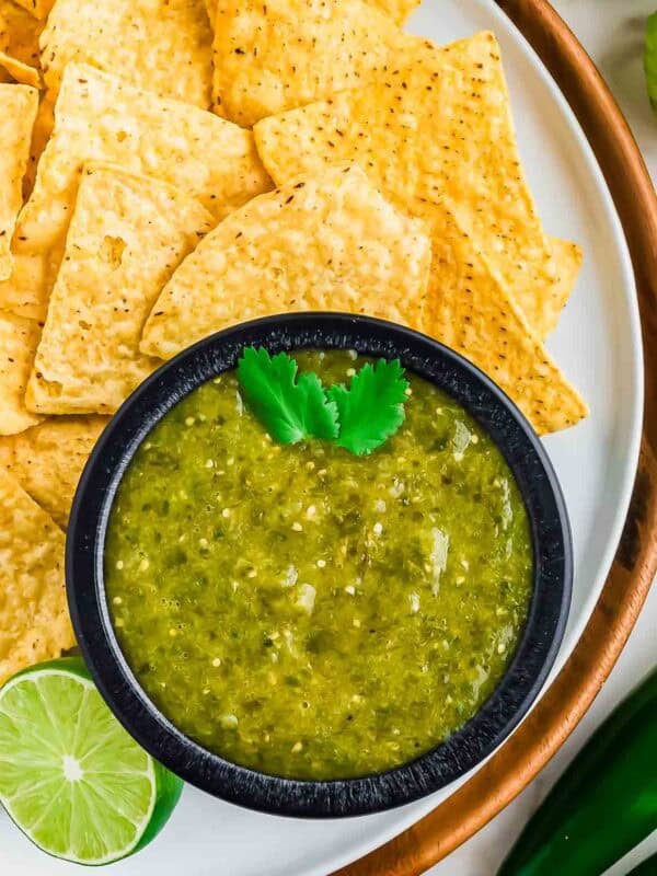 A black bowl filled with salsa verde garnished with cilantro, surrounded by yellow tortilla chips on a white plate. A lime slice and part of a jalapeño are also visible.