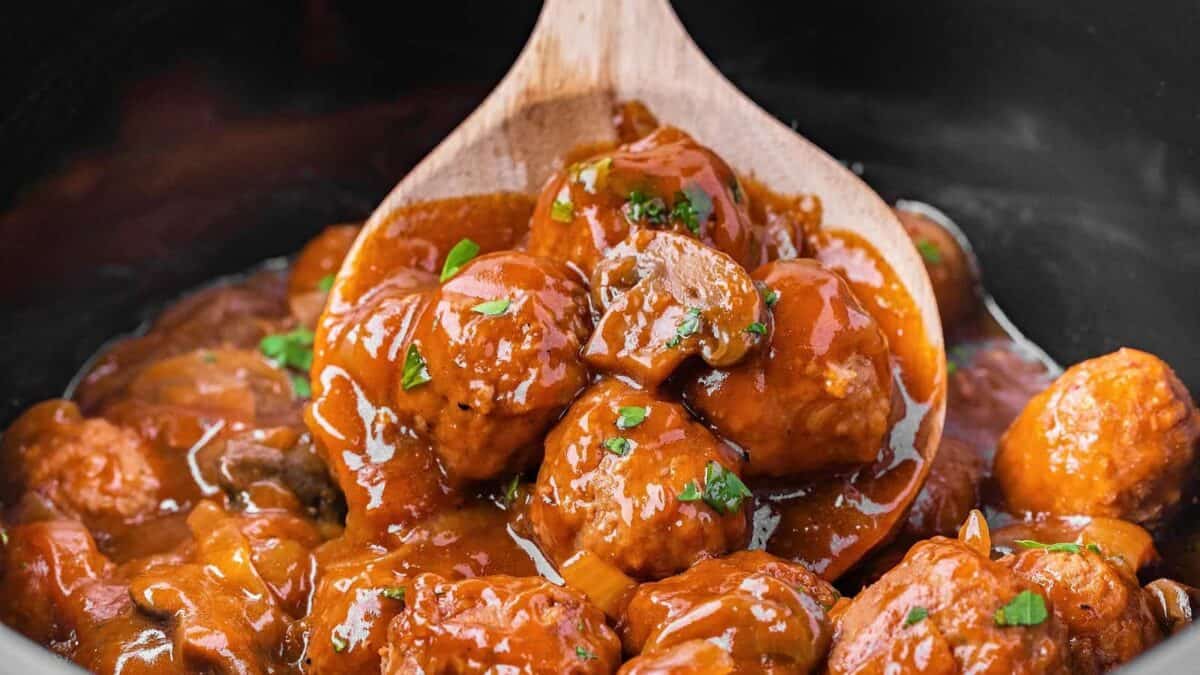 A wooden spoon lifts saucy meatballs garnished with chopped parsley from a pot filled with rich, red tomato sauce.