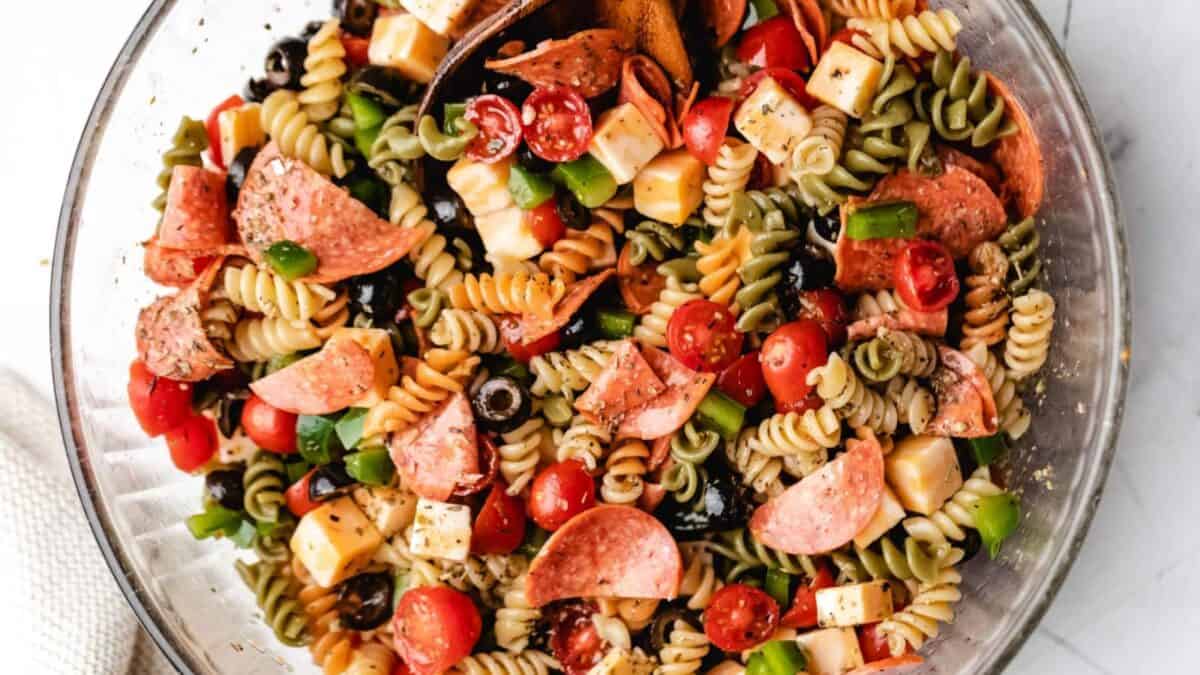 A glass bowl filled with colorful pasta salad made of rotini noodles, pepperoni slices, cherry tomatoes, green bell peppers, black olives, and cheese cubes, all mixed with seasoning.