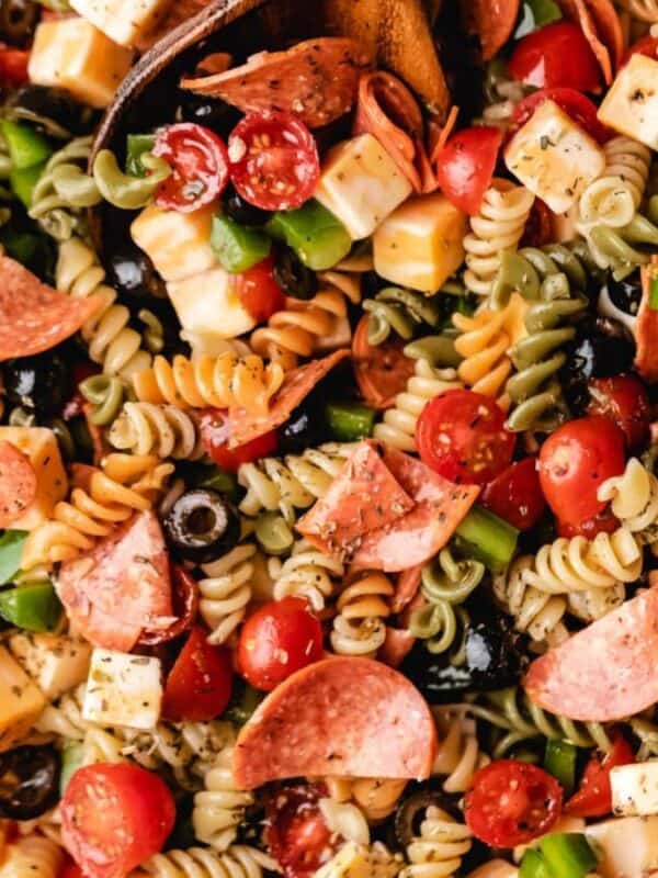 A glass bowl filled with colorful pasta salad made of rotini noodles, pepperoni slices, cherry tomatoes, green bell peppers, black olives, and cheese cubes, all mixed with seasoning.
