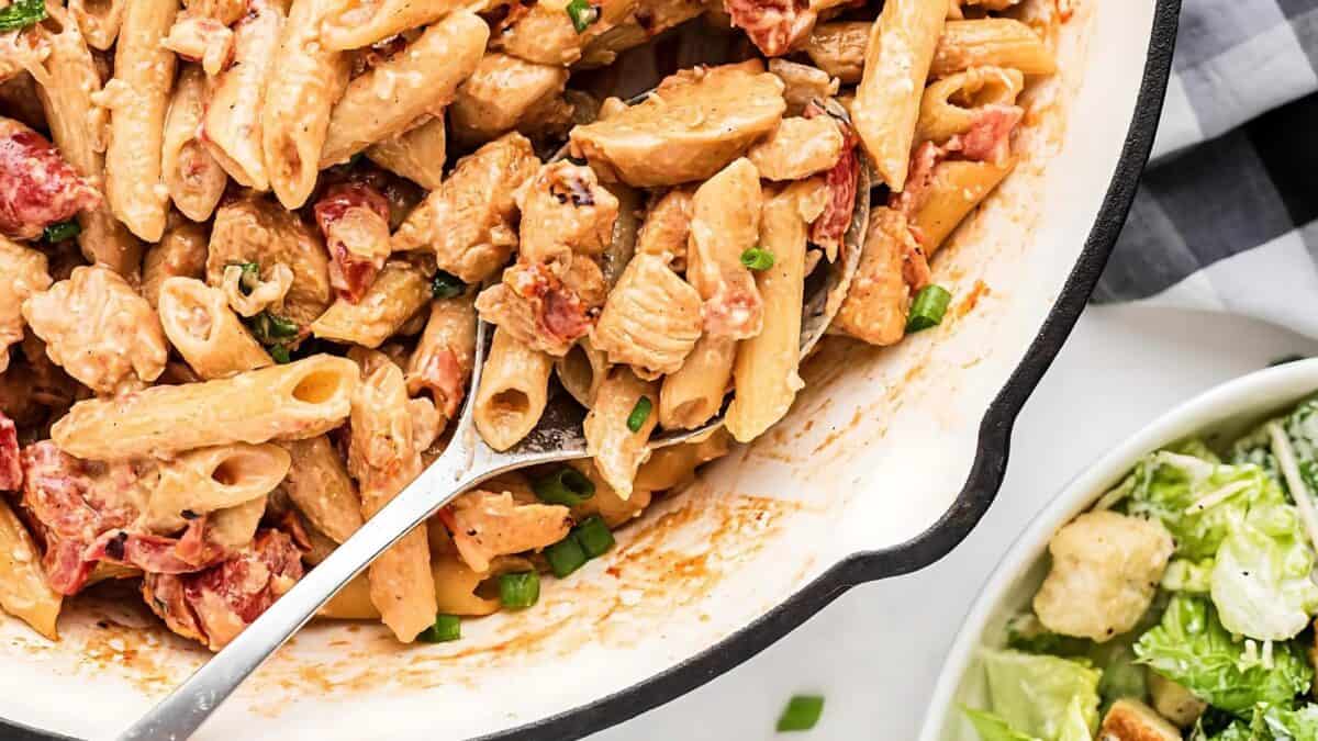 A serving spoon scoops creamy penne pasta with chicken and diced tomatoes from a white dish, next to a bowl of Caesar salad with croutons on a table.