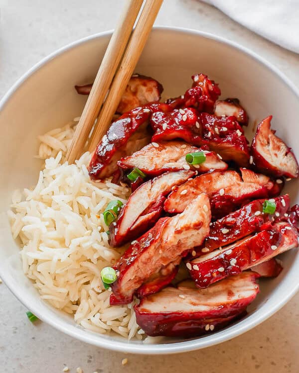 A white bowl filled with white rice and sliced char siu chicken breasts, garnished with chopped green onions and sesame seeds, with chopsticks resting on top. Small bowls of sesame seeds and green onions are nearby.