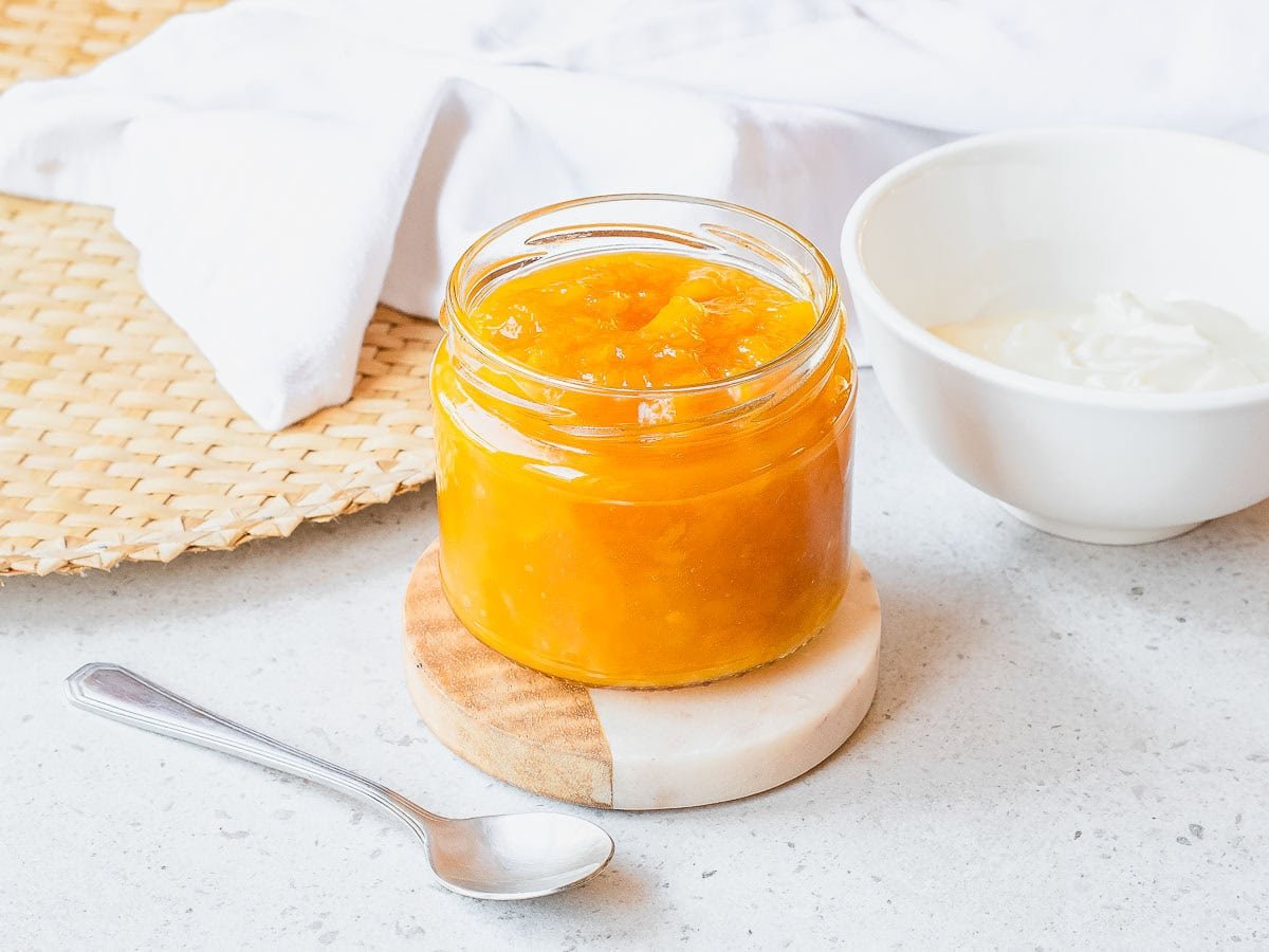A glass jar filled with bright orange mango jam sits on a small round wooden coaster, next to a silver spoon and a white bowl, on a light countertop with a woven placemat and white cloth in the background.
