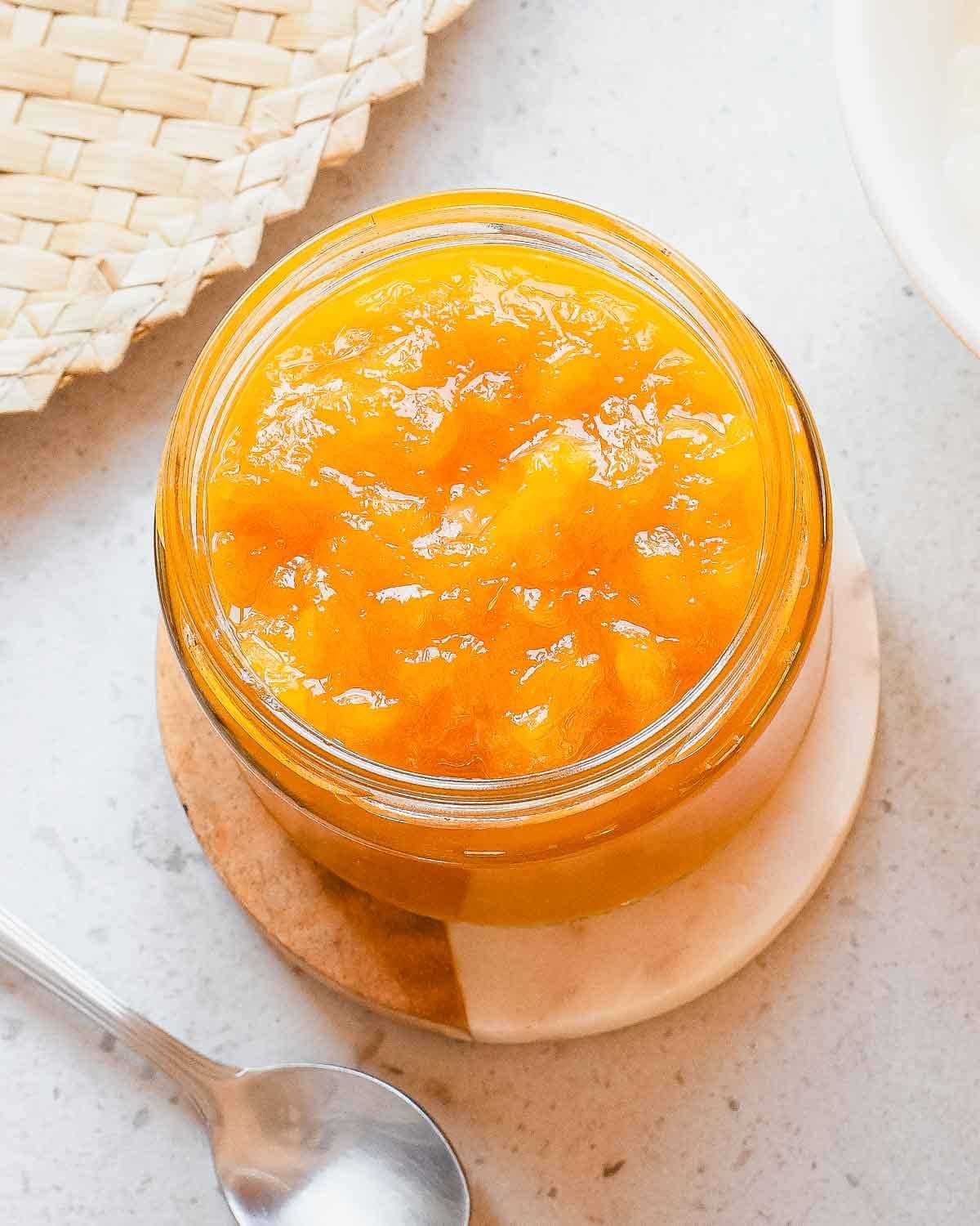 A glass jar filled with bright orange homemade mango jam sits on a round coaster, next to a spoon and a woven placemat on a light-colored surface.