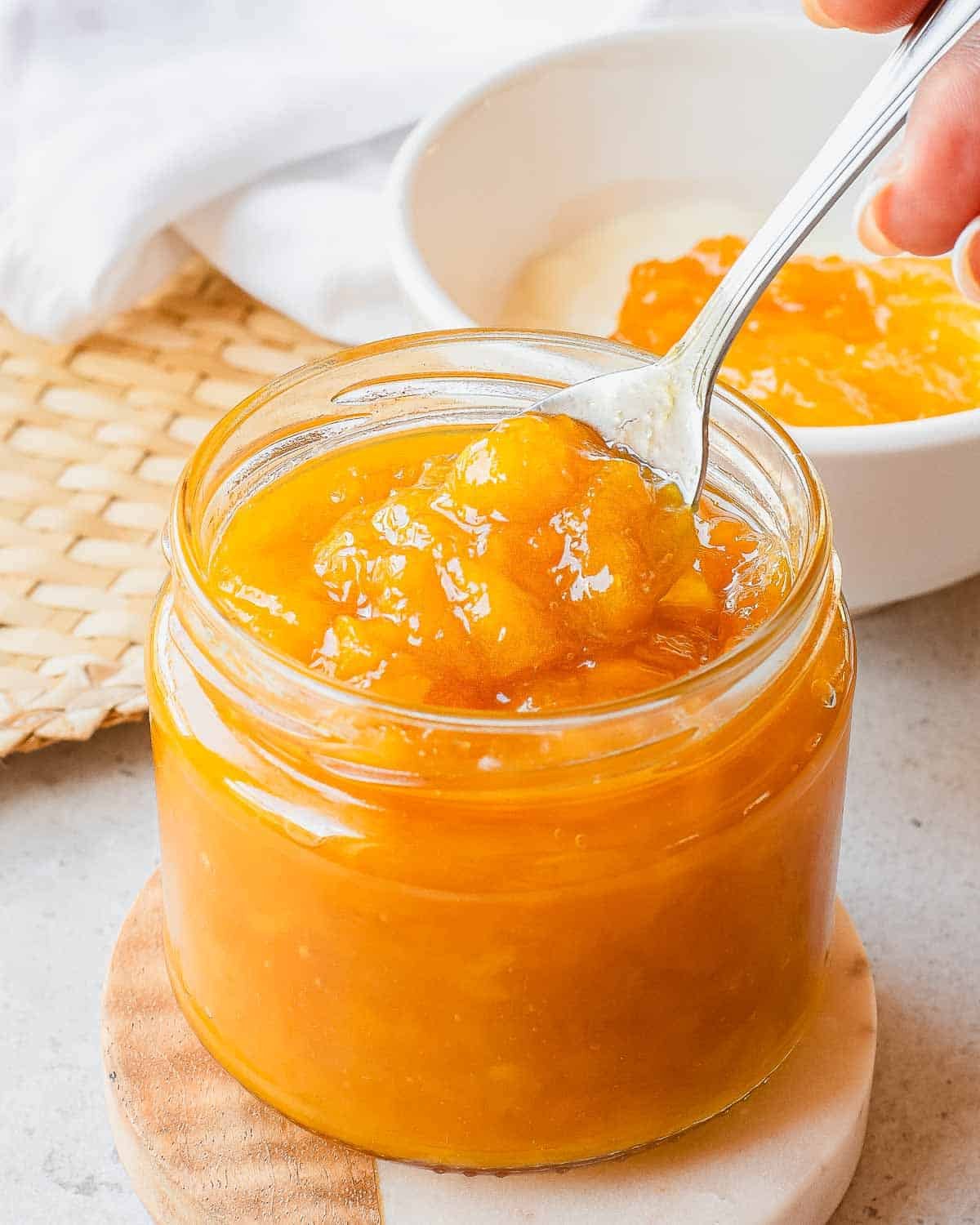 A close-up of a glass jar filled with bright orange homemade mango jam, with a spoon scooping some out. In the background, a bowl with more jam is partially visible on a light surface.