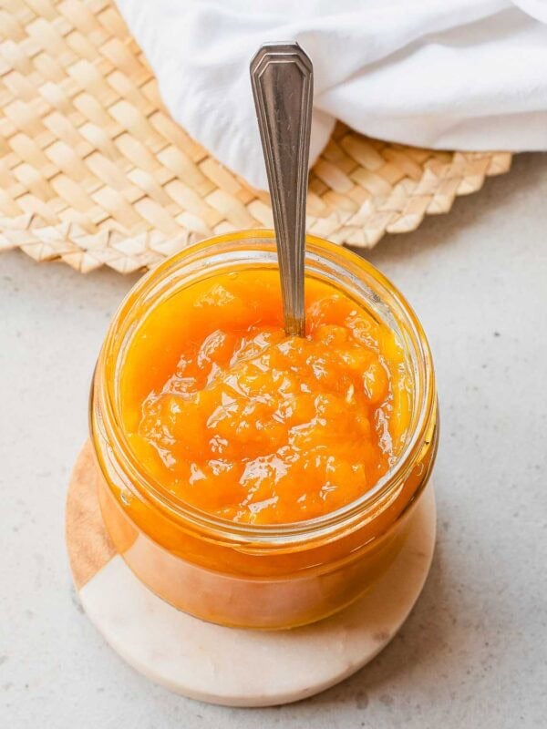 A glass jar filled with bright orange mango jam sits on a round coaster, with a metal spoon inside. A woven placemat and a white cloth are in the background.
