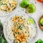 A plate of white rice topped with a cheesy green salsa chicken bake, garnished with chopped herbs and served with a lime wedge. Fresh tomatillos, a tomato, and cilantro are nearby on the table.