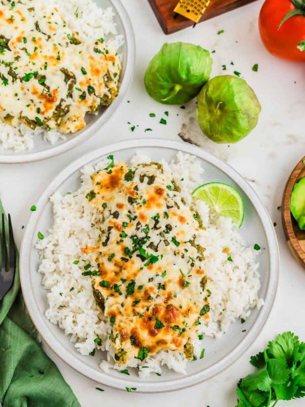 A plate of white rice topped with a cheesy green salsa chicken bake, garnished with chopped herbs and served with a lime wedge. Fresh tomatillos, a tomato, and cilantro are nearby on the table.