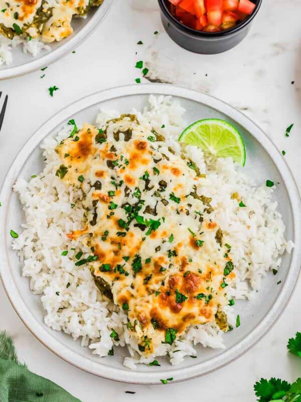 A plate of white rice topped with cheesy chicken and green salsa sauce with herbs, garnished with a lime wedge. Sliced avocado, chopped tomatoes, cilantro, and forks are nearby on a white tabletop.