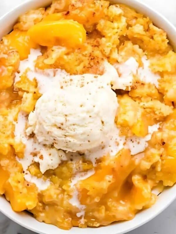 A bowl of peach cobbler topped with a scoop of vanilla ice cream, surrounded by fresh peaches on a white surface with a spoon and fork nearby.