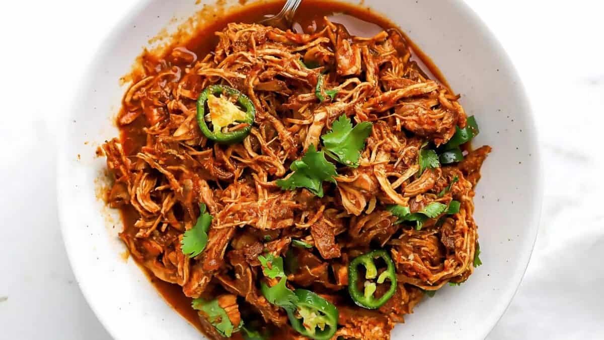 A bowl of shredded chicken in a rich red sauce, garnished with sliced jalapeños and fresh cilantro. A fork is partially visible in the bowl, and everything sits on a white background.