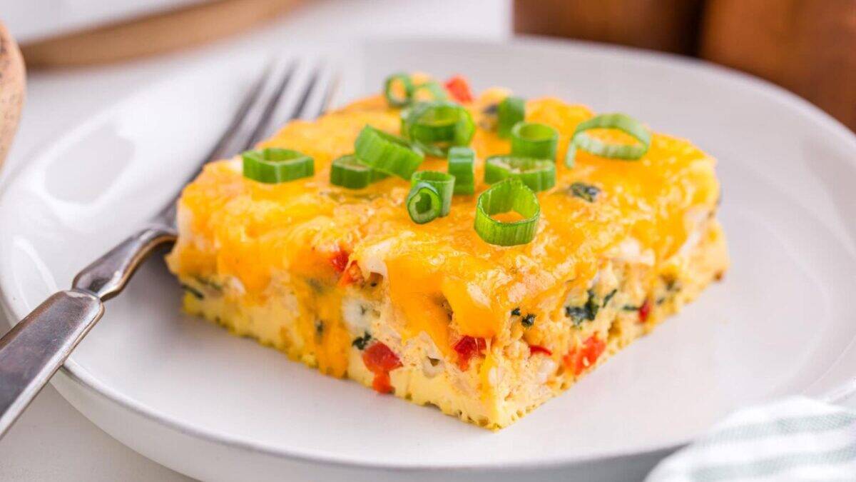 A square slice of cheesy egg casserole with chopped green onions on top, served on a white plate with a fork. The casserole contains visible bits of red and green vegetables.